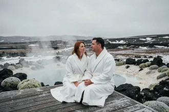 Couple en robes blanches assis sur une terrasse en bois au bord d'une piscine géothermique en Islande, entouré de roches volcaniques noires et de brume naturelle