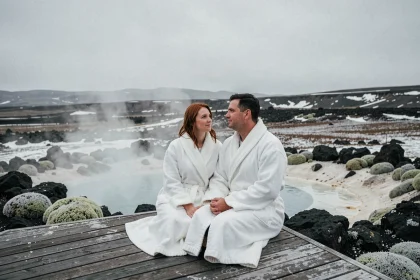 Couple en robes blanches assis sur une terrasse en bois au bord d'une piscine géothermique en Islande, entouré de roches volcaniques noires et de brume naturelle